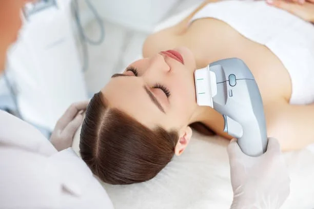 Person receiving facial treatment with handheld device in Anchorage spa, operated by professional; both wear white attire.