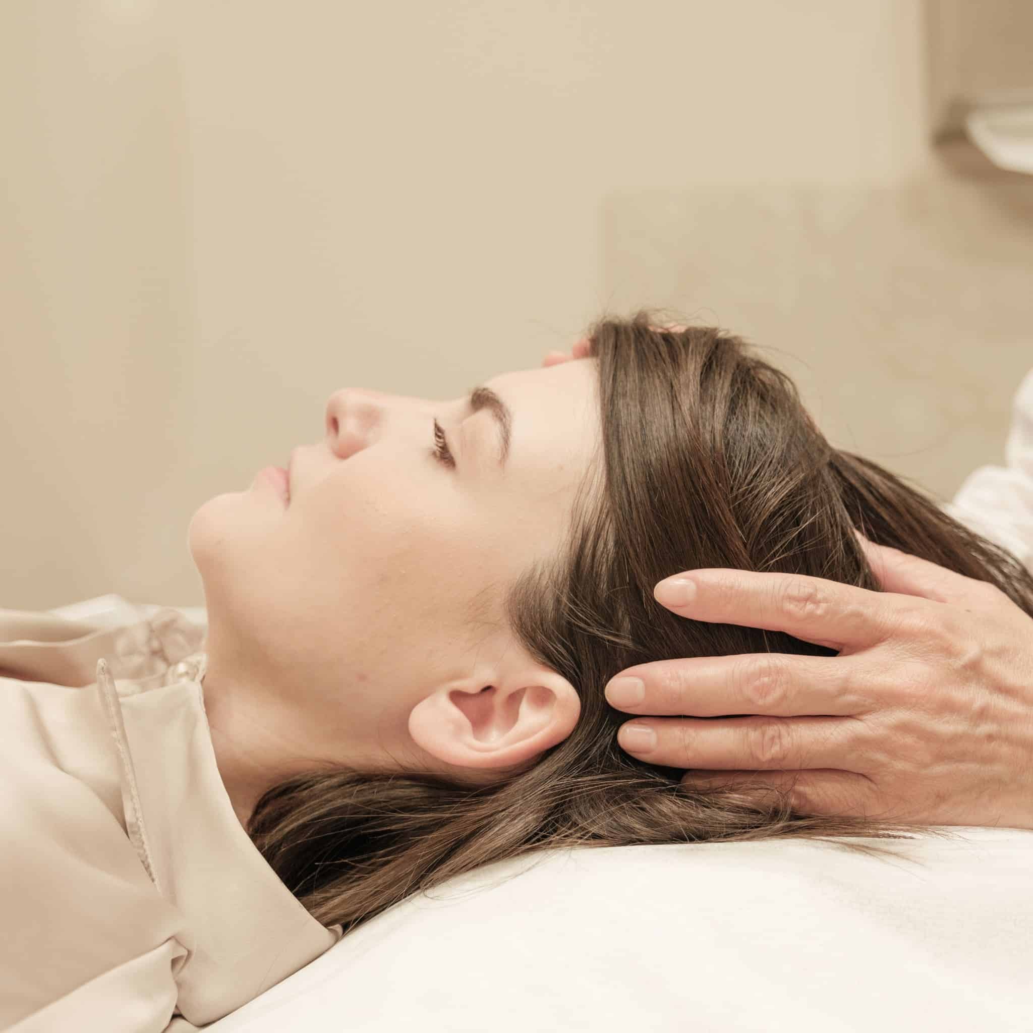 A person reclines as another gently supports their head, indicating a relaxation or skin rejuvenation session in a neutral room.