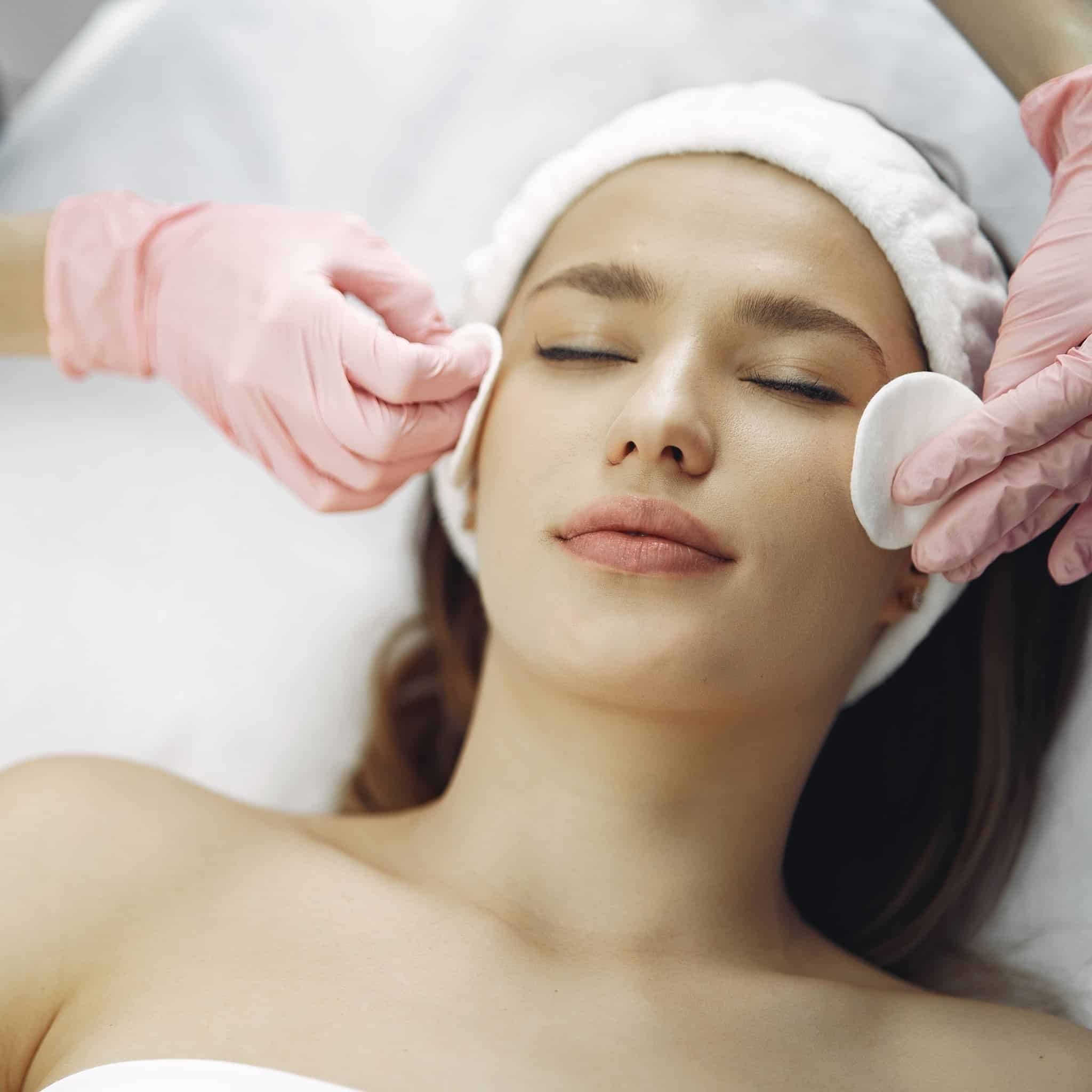 A woman with closed eyes gets a facial while someone in pink gloves places cotton pads on her face.