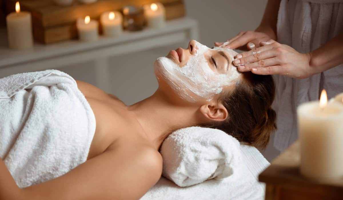 Person with a facial mask relaxing on a spa table as another gives a forehead massage, candles glowing in the background.