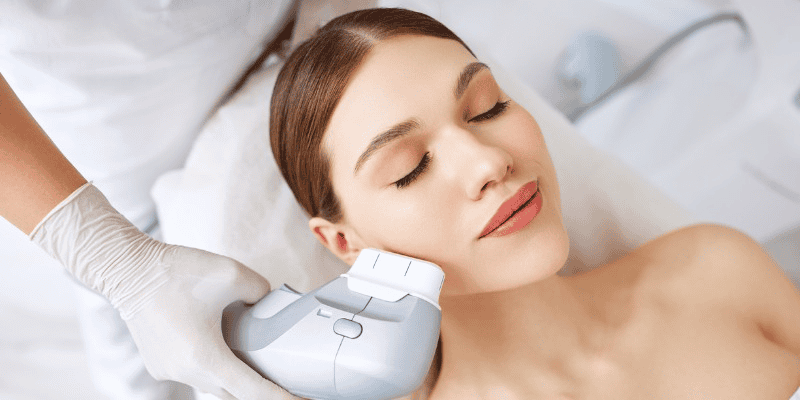 A woman with closed eyes gets a facial treatment using a handheld device by a professional wearing gloves.