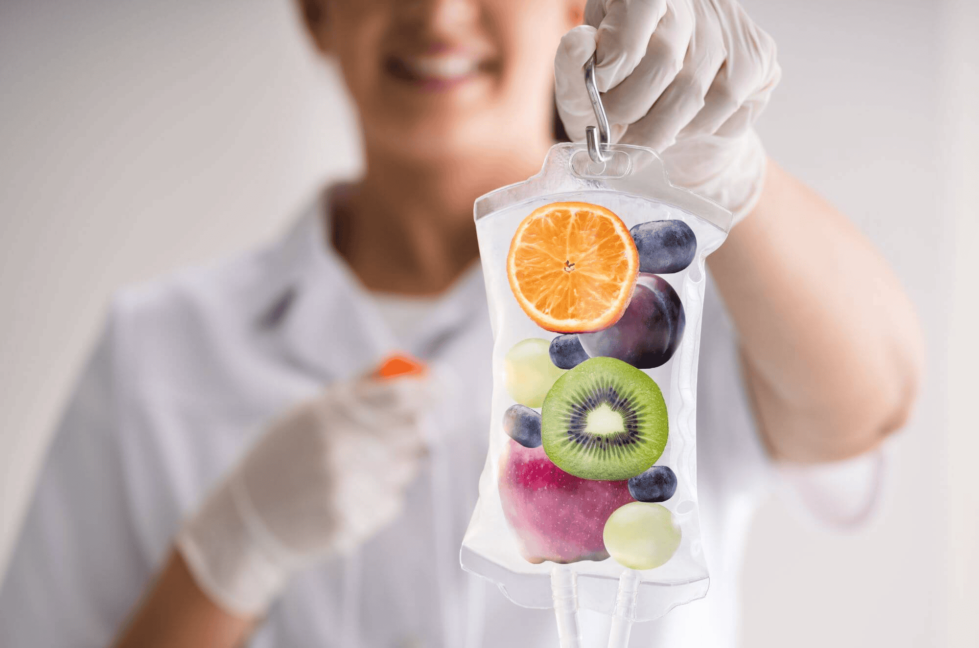 Gloved hands hold an IV bag filled with fruits like orange, kiwi, apple, grapes, and blueberries.