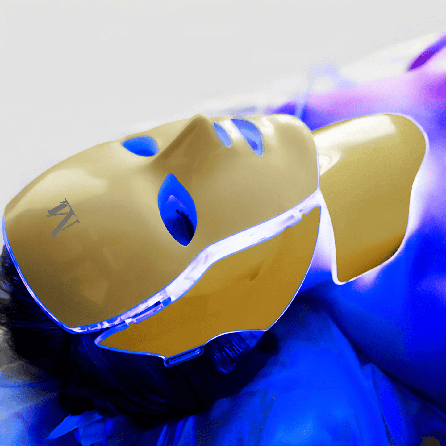 A person relaxes wearing a gold LED light therapy mask under blue and purple lighting.