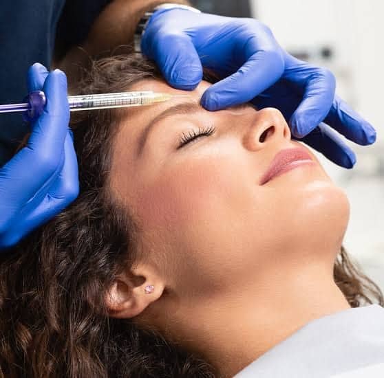 Someone in blue gloves injects the forehead of a person with closed eyes.